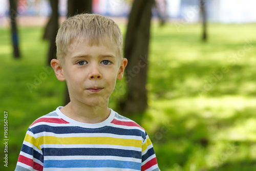 Portrait of a fair-haired young boy in a colorful striped shirt standing against a blurred green park background.
