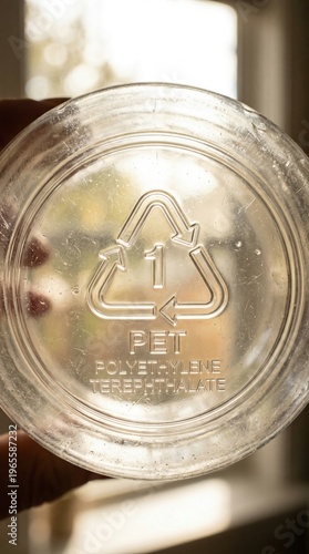 A hand holds a transparent plastic bottle with a recycling symbol prominently displayed. The sun creates a warm glow, illuminating the bottle's surface while soft shadows dance around it.