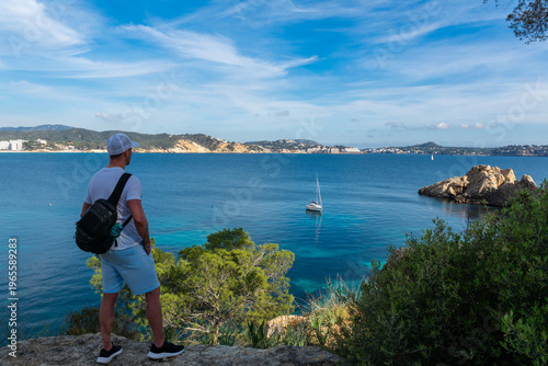 Man with backpack looking at turquoise sea and sailboat from coastal viewpoint in Mallorca