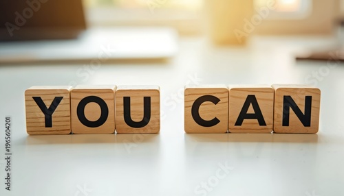 Wooden blocks with letters spell out YOU CAN on a white desk. This image promotes motivation and belief in ones ability to achieve goals. Positive affirmation for success and personal growth.