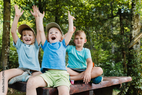 Photos Three joyful children sitting on a picnic table raising their arms in celebration at a forest camp