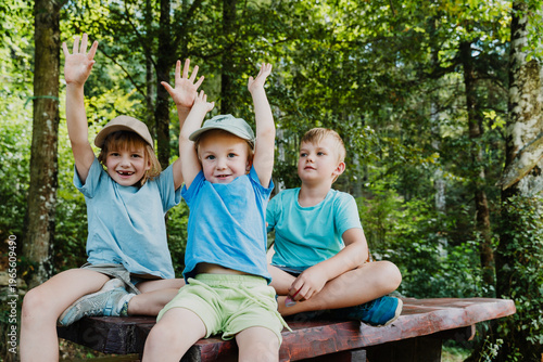 Εκτύπωση καμβά Three joyful children sitting on a picnic table raising their arms in celebration at a forest camp