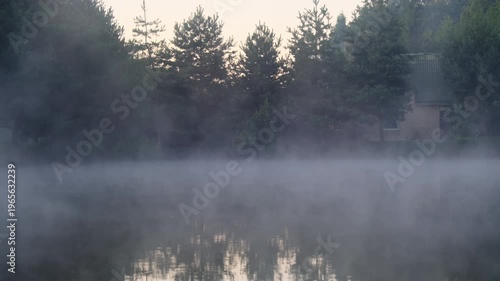Haze flows rise over lake surface against cottage in fir forest. Tranquil morning scene with mysterious mist hiding river and pine wood slow motion