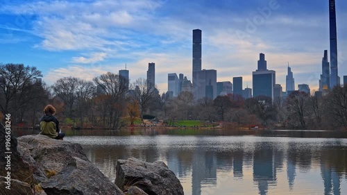 Rear view of a person sitting on the boulder near the pond in Central Park, New York, USA. Manhattan skyline against cloudy sky in the backdrop.