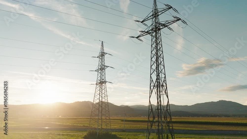 Power lines and towers are visible in a rural area as the sun sets in the background, highlighting energy infrastructure in nature.