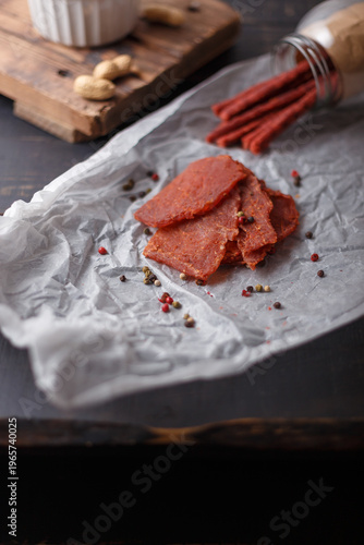 Dry pork meat slices on white paper. Fish sticks, salmon jerky in jar on black wooden background. Bowl of unpeeled peanuts. Vertical shot