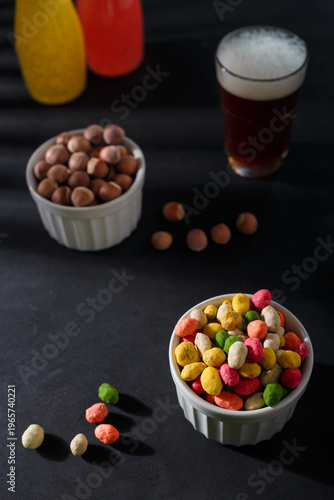 Multiflavored peanuts in bright multicolored icing in white bowl on black table background. Beer and non alcohol drinks, hazelnuts. Vertical shot