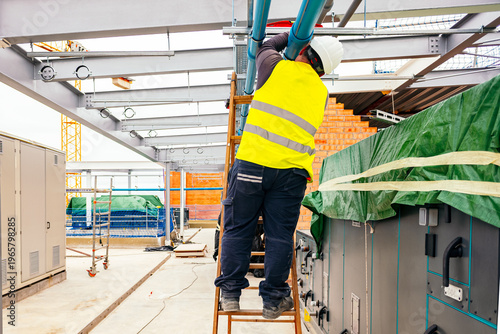 Construction worker wearing safety vest and helmet, standing on a wooden ladder, installing pipes on a building rooftop, with construction equipment and materials in the background.