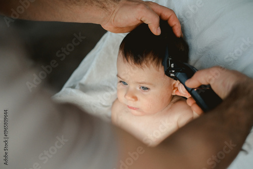Soft light glows on a tender moment. Father's hand rests gently on a baby's head. The infant gazes calmly, fingers near lips. The baby's eyes hold quiet curiosity and trust