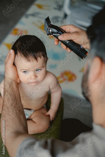 Soft light glows on a toddler's curious face. Parent gently holds baby's head while trimming hair. Baby looks at camera with wide, trusting eyes. Moments of tenderness during a simple, everyday task