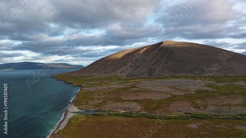 Aerial view drift tracks the shoreline and cliffs with Gilmimyl rising ahead. Summer morning clouds hang low above Chukotka, Russia and Gilmimyul Bay. Wide landscape for tourism media