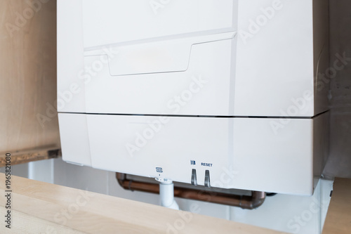 Detailed view of a newly fitted gas central heating boiler seen located in a kitchen cupboard. Part of the plumbing, water drain pipe and thermostat dial is visible.