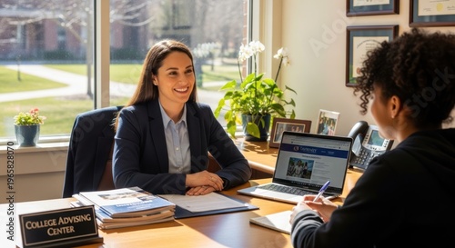 A college advisor smiles warmly as she engages in a counseling session with a student. The advisor, a young woman with dark hair, sits at a wooden desk in a college advising center.