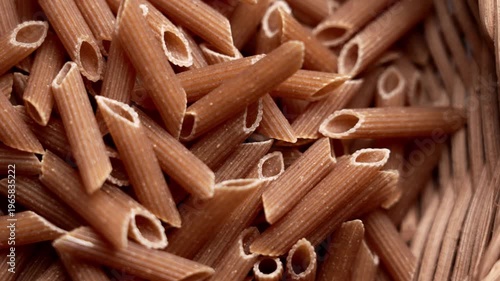 Dry dark wheat penne rigate pasta inside a traditional wicker basket. Wholesome uncooked ingredient. Top view. Rotation
