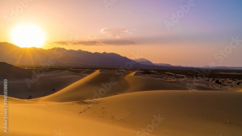Desert dunes bask in warm light of setting sun, mountains in distance, colorful sky