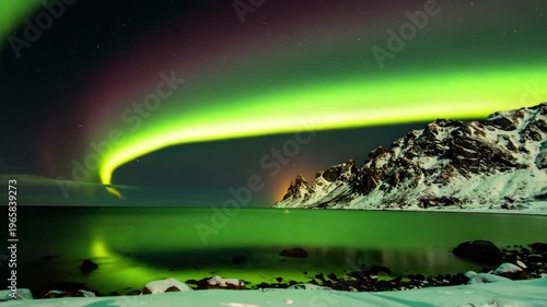 Dramatic green aurora borealis dances above snow-capped mountains and tranquil sea at night