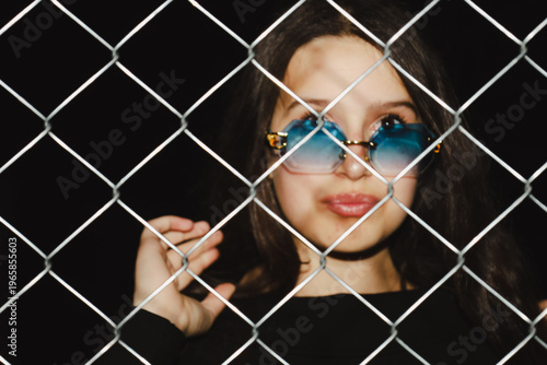 Φωτογραφία A young girl's portrait in stylish blue sunglasses viewed through a metal chain-link fence
