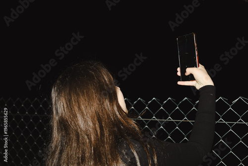 A woman with long brown hair holds a smartphone in front of her, posing against a chain-link fence at night. Harsh flash light focuses on her hand and hair texture, leaving the background in deep dark