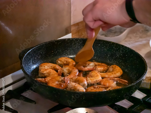 Man frying shrimp in a pan on gas stove. Process of cooking seafood with wooden spatula at home. Culinary preparation of healthy dish. Home lifestyle, kitchen leisure, nutrition concept.