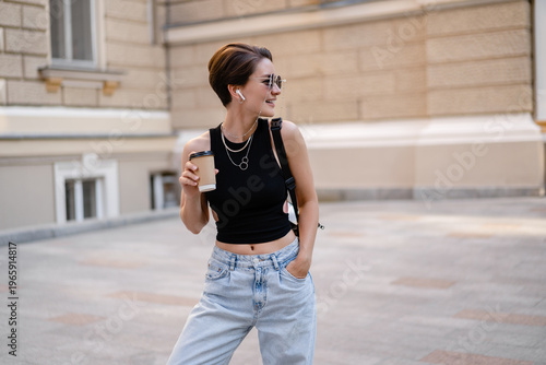 stylish woman with short haircut walking in street with coffee wearing black top, jeans and sunglasses accessories