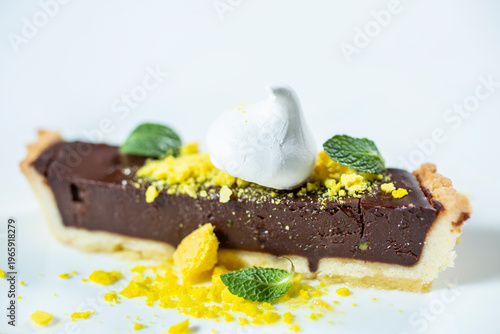 A piece of chocolate tart with a light base, decorated with white icing, mint leaves and yellow sprinkles on a white background.