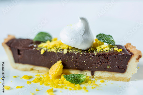A piece of chocolate tart with a light base, decorated with white icing, mint leaves and yellow sprinkles on a white background.