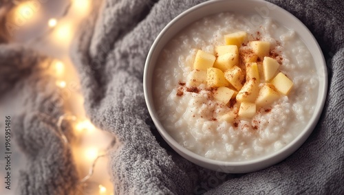 Warm homemade rice pudding with diced apples and cinnamon, served in a white bowl on a soft grey blanket with bokeh lights.