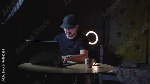 A man in black clothes sits at a table. He wears a baseball cap and glasses. He works remotely. Freelancer. Night. The lights are on. There's a laptop on the table. A ceramic mug. Home interior. Stora