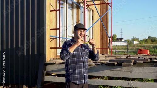 An adult man in front of a house under construction. He wears a dark shirt and protective glasses. It's warm weather. A wooden house.