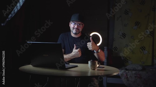 A man in black clothes sits at a table. He wears a baseball cap and glasses. He works remotely. Freelancer. Night. The lights are on. There's a laptop on the table. A ceramic mug. Home interior. Stora