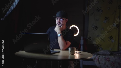 An adult man sitting at a table. Working on a laptop in a dark room. Lighting equipment. Film studio. Props in the room. Warehouse. Dressed in dark clothes. Adult. Bearded. Wearing glasses.