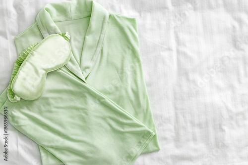 Top view of folded pastel green woman pajama with satin sleep eye mask on white bed. Minimal flat lay cozy comfort pyjamas for sleeping and relaxed. Aesthetic lifestyle photo of stylish sleepwear