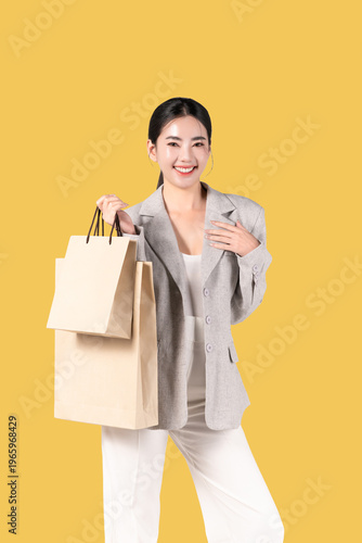 Happy young Asian woman holding shopping bags on yellow background, for discount clothes and mid year sale concept. women glamour lifestyle in holiday.