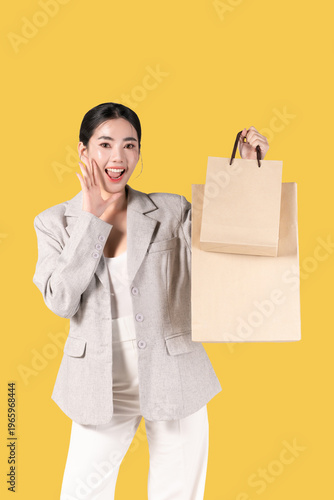 Happy young Asian woman holding shopping bags on yellow background, for discount clothes and mid year sale concept. women glamour lifestyle in holiday.