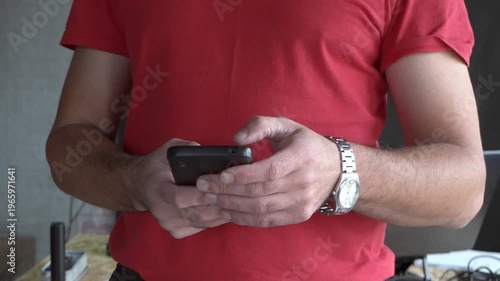 A man holding a smartphone. Holding a phone in his hand. Red T-shirt. Close-up. Writing a message.