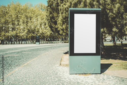 Blank outdoor billboard mockup on curved cobblestone road near park trees, vertical advertising display template with copy space in sunny urban landscape