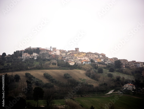 Borgo medievale italiano arroccato su una collina, circondato da campi verdi, ulivi e dolci colline. Le case in pietra con tetti in coppi rossi si raccolgono attorno alla torre storica del centro