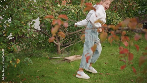 White girl dancing in backyard garden, playful twirls and small cartwheel attempt among autumn leaves casual hoodie and jeans, white sneakers on green grass, sunlight through branches, raised planter
