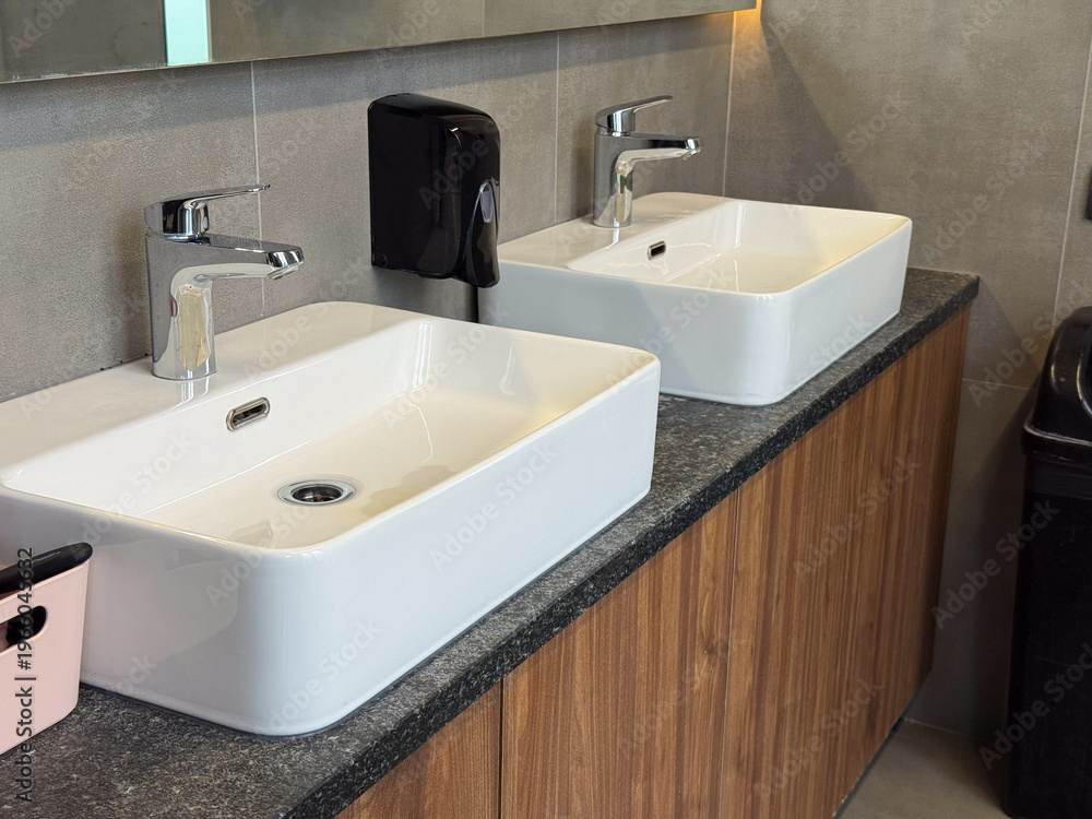 Fototapeta premium Double ceramic sinks with chrome faucets installed on stone countertop in shared restroom. Hygiene routine, simultaneous usage, public facility design and sanitation efficiency concept