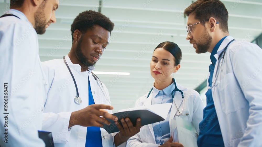 Fototapeta premium Focused male doctor holding chart while colleagues gathering around him in bright hospital hall. Reviewing patient notes. Clarifying duties. Coordinating tasks for busy clinical shift.