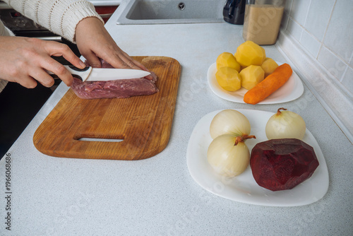Person slicing raw meat on a wooden cutting board, with fresh ingredients like potatoes, carrots, onions, and beetroot prepared for cooking borscht