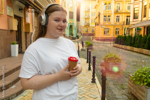 Young woman in a white t-shirt wearing wireless headphones and holding a paper coffee cup while standing on a beautiful historical city street