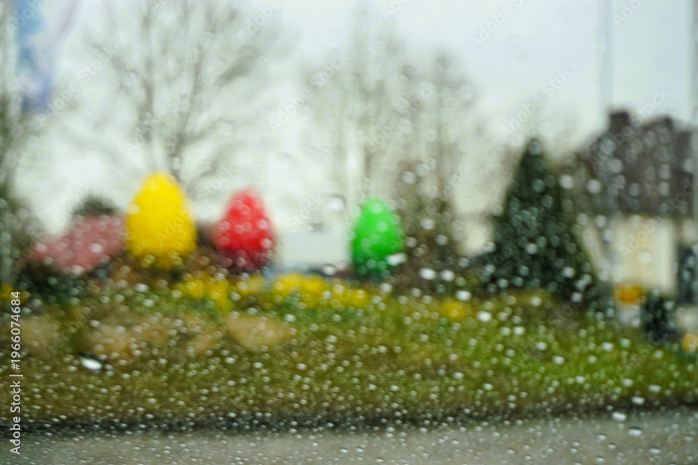 Obraz premium Abstraktes Osterpanorama mit Regentropfenmuster vor Glasscheibe vor bepflanztem Erdhügel mit grünen Narzissen, Gras, Tannen und bunten großen Osterei-Figuren vor Himmel bei Schnee, Sturm im Frühling