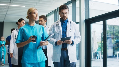 Focused nurse holding coffee and clipboard while walking with calm expression through clinic hallway. Male doctor beside her reviewing notes on tablet and preparing tasks. Team moving forward.