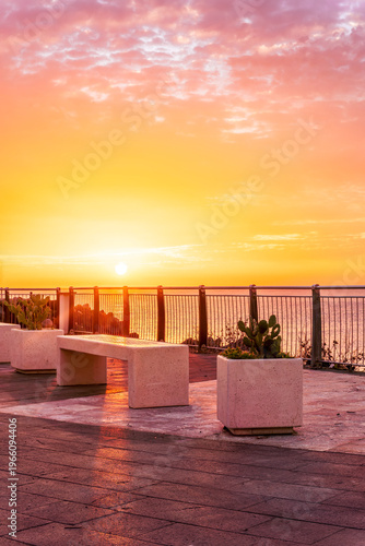beautiful landscape with white stone bench on seafront embarkment during beautiful sunrise or sunset with sidewalk with pavement, sea surf and nice cloudy sunset sky