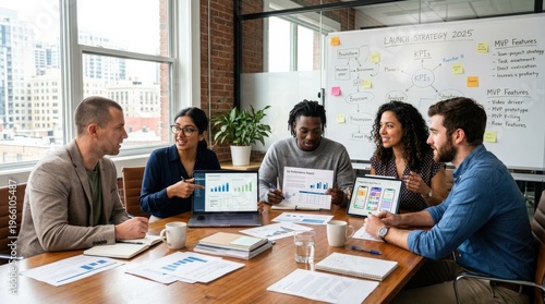 A focused entrepreneurial team seated around a conference table in a bright startup meeting room, some reviewing printed statistical reports while others gesture at laptop screens and tablets