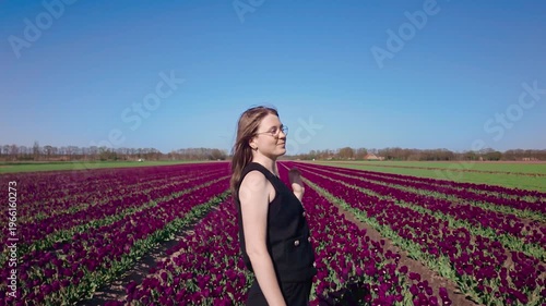 Woman stands among blooming tulip field under clear blue sky, looking around and enjoying spring landscape, peaceful outdoor moment, floral lifestyle scene, seasonal beauty.