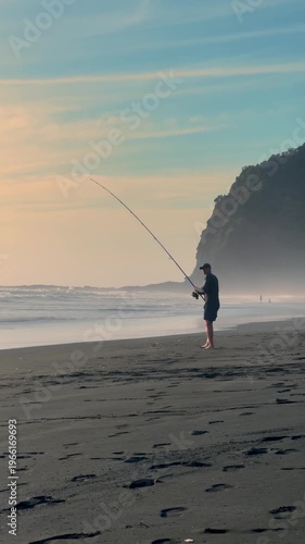 A lone fisherman casts his line into the surf at Karekare Beach, New Zealand. He hopes to catch fish for dinner, while enjoying the peace of the ocean at sunset.