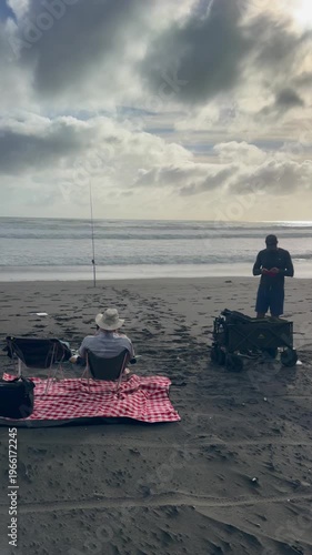 Two men enjoy a day of fishing on the beach in Karekare, Auckland, New Zealand. One man sits and relaxes while the other prepares the fishing gear near a wagon.