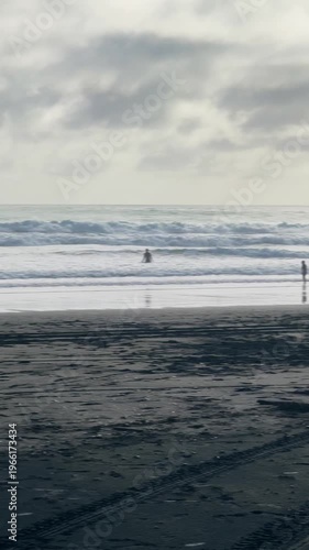 People enjoy the ocean waves at Karekare Beach in Auckland, New Zealand. Beachgoers are swimming and playing in the water on a cloudy day at the popular beach.